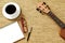 Top view of ukulele, notebook, pen, coffee cup and glasses on brown background