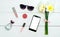 Top view on white wooden background with lip balm, lip gloss, perfume, sunglasses and phone with white screen around bouquet.