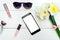 Top view on white wooden background with lip gloss, perfume, sunglasses and phone with white screen around bouquet of narcissus.