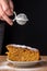 Top view of woman`s hand that is going to pour sugar over portion of carrot cake, in blue and white plate on wooden table with bl