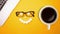 Top view of work place desk, laptop with glasses , cup of black coffee and smiley face on empty bright yellow background