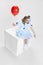 Top view of young girl wearing Halloween dress of dancing clown with spooky make-up sitting on box with red balloon