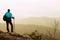 Tourist in blue green windcheater with sporty backpack and poles in hands stand on rocky view point and watching into deep valley