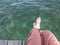 A tourist in brown pants sitting on the wooden bridge hanging legs with barefoot to the crystal clear water in the sea