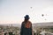 A tourist girl in a hat admires hot air balloons flying in the sky over Cappadocia in Turkey. Impressive sight.