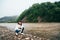 Tourist girl in jeans and white sweatshirt sitting on the bank of the dirty mountain river surrounded by forest