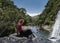 Tourist girl sitting on the rock and looking down at the edge of waterfall