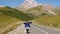 A tourist girl in a windbreaker walks along an asphalt road crossed by cows. View from the back. In the background are