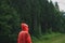 Tourist hiker in red raincoat stands in rain in mountains against coniferous forest background and looks away. Minimalistic photo