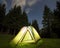 Tourist hikers tent brightly lit from inside on green grassy forest clearing among tall pine trees under dark blue starry sky on