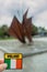 Tourist holding badge with National flag and sign Ireland in focus, Galway Hooker monument in Eyre Square out of focus