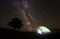 Tourist illuminated tent near big tree, camping in mountains at night under starry sky