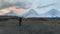 A tourist with a large backpack walks along the tundra against the backdrop of three majestic volcanoes.