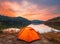 tourist lit tent by the lake at sunset. Dramatic sky. Orange lit inside the tent and a fire over the misty river at