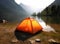 tourist lit tent by the lake at sunset. Dramatic sky. Orange lit inside the tent and a fire over the misty river at