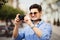 Tourist Man With Camera Taking Photos On Street. Portrait Of Handsome Smiling Male Holding Camera, Making Photo Of Interesting Pla