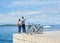 Tourist pair, man and woman with bicycles on high paved stone sidewalk near sea water on sunny day