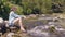 Tourist woman sitting on shore of stony river and resting while summer hike