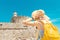 Tourist woman on vacation posing with hat and backpack in front of the old ruined fort and lighthouse in Rhodes, Greece