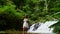 Tourist Woman in White Dress Walking Barefoot near Cascade Tropical Waterfall