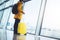 Tourist with yellow suitcase backpack is standing at airport on background large window, traveler person waiting in departure loun