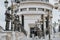 Tourists exploring historic statues in urban square with ornate lampposts.