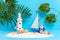 Toy sailing boat, captain on sandy island shore with lighthouse, monstera leaves and palm tree, seashells and starfish