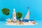 Toy sailing boat, captain on sandy island shore with lighthouse, monstera palm tree, coral, seashells and message bottle