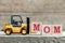 Toy yellow forklift hold letter block M to complete word mom on wood background (Concept for mother 's day