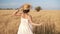 Tracking footage of a beautiful girl in white summer dress and straw hat running freely by wheat field. Backside view