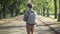 Tracking shot of confident intelligent teenage boy with backpack walking in sunshine in summer park. Back view of