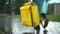 Tracking shot of yellow delivery bag in hand of unrecognizable courier in rain coat walking in urban park. Young