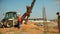 Tractor lifts a steel rod and put it in a hole in the ground. Construction workers are helping him.