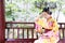 Traditional Asian Japanese beautiful Geisha woman wears kimono hold a fan sit on a bench in a summer nature garden