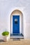 Traditional blue door and whitewashed walls of Greek house entrance and flower pot with rich foliage. Midday, summer