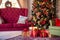 Traditional festive advent interior decoration: living room with maroon sofa, armchair and Christmas tree and gift boxes under it