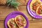 Traditional Mexican tacos with meat and vegetables in purple paper plates served for takeaway on wood table with pineapple