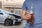Traffic accident and alcohol concept. Drunk driver is holding bottle with alcohol in hand. Crashed car in background.