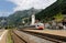 A train travels on the railway curving through a Swiss village with churches on the hillside and majestic Alpine mountains