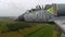 A training gray blue jet plane flies and move towards the goal over green yellow fields, forest and a river against a dark sky