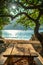 Tranquil Beach Scene with Wooden Picnic Table Under Shade Trees and Sparkling Sea in the Background