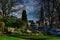 Tranquil urban park scene with benches and lush greenery, set against a backdrop of historic buildings and blue sky with wispy