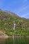 The tranquil waters of Trollfjorden reflect the sunlit, verdant landscape and a thin waterfall descending