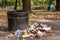 trash can overflowing in busy city park, with cigarette and litter strewn about