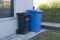 Trash cans of black and blue on wheels stand near the wall. Dirty mobile trash bins and household waste. Sorting garbage into