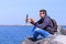 Travel blogger man shoots video on smartphone sits on stone at sea beach.