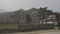 A traveler with a backpack walks against the background of an old house with a fence and mountains with vineyards. Sunny