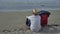 Traveler man in a hat sits by the sea on the sand next to a red suitcase, watches the waves