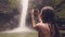 Traveler woman using smartphone for photo splashing waterfall in jungle forest. Woman tourist shooting video tropical