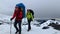 Travelers with large backpacks and trekking poles walk along a snowy trail.